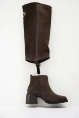 Bota de caña alta confeccionada en cuero gamuzado color marrón, con diseño de polaina desmontable que permite versatilidad en el estilo. Presenta una puntera redondeada y un taco cuadrado de altura media, ofreciendo un look clásico con un toque moderno.