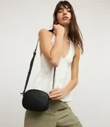 Cartera tipo camera bag pequeña, color marrón camel, de forma ovalada con superficie texturizada. Cuenta con correa ajustable larga, cierre principal con cremallera dorada y un pequeño bolsillo frontal con cremallera.