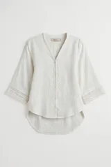 Camisa de lino color blanco, con escote en V, mangas tres cuartos con detalles de encaje en los puños y ruedo asimétrico más largo en la parte trasera.