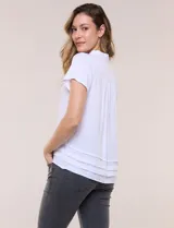 Camisa blanca de rayón con cuello camisero, escote en V y mangas cortas. Presenta abotonadura frontal con botones nacarados y detalles de tablas decorativas en el ruedo.
