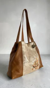 Cartera tipo shopper de cuero, con diseño combinado en colores caramelo y perla, asas de hombro y arte pintado a mano en el frente.
