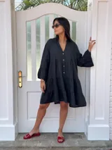 Vestido corto de lino color negro, con escote en V y botones frontales de coco. Presenta mangas largas con volados y un corte amplio con frunces en el torso y ruedo.