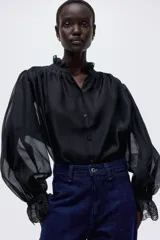 Blusa negra de tejido transparente con cuello alto y puños con volado y detalles de broderie. Presenta frunces en los hombros y cierre frontal con botones.