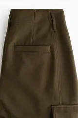 Pantalón cargo de sarga color verde militar, con tiro alto, cierre frontal con botón y corte holgado en las piernas estilo barrel. Cuenta con bolsillos laterales, bolsillos de fuelle con solapa en los muslos y trabillas abotonadas en los tobillos.
