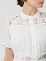 Vestido midi blanco de lino orgánico y viscosa, con cuello mao, mangas cortas y cierre frontal de botones. Presenta detalles de encaje en los hombros, el pecho y la cintura.