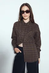 Camisa tipo leñadora de corte oversize, con estampado de cuadros en tonos negro y marrón. Presenta cuello clásico, cierre frontal con botones y mangas largas con puños abotonados.