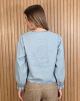 Camisa de denim de algodón ligero, con cuello en V adornado con grandes volados frontales y cierre de botones. Presenta mangas largas con puños elásticos fruncidos.