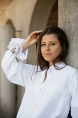 Camisa de lino blanca con cuello camisero, cierre frontal de botones de nácar y mangas largas con puños en contraste de tela rayada.