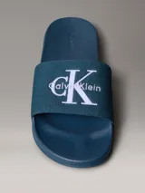 Chinelas de color azul con banda superior de material textil que presenta el logotipo de Calvin Klein bordado en blanco. La suela es ergonómica y antideslizante, con el nombre de la marca grabado en el lateral.