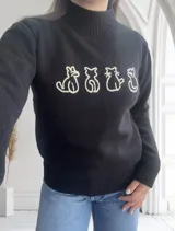 Sweater negro de tejido de punto con cuello alto y diseño de cuatro gatos blancos en el frente.