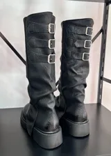 Botas negras de caña alta con suela track y tres hebillas decorativas en la parte trasera.