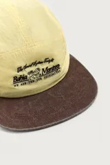 Gorra de 5 paneles de color amarillo claro con tejido ripstop, visera plana marrón de efecto desgastado y bordado frontal en negro. Cuenta con ajuste en la parte posterior.