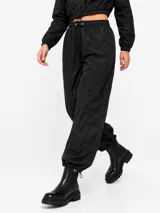 Pantalón estilo parachute negro, confeccionado en tejido liviano con acabado ligeramente brillante. Cuenta con cintura elástica con cordón ajustable y puños fruncidos en los tobillos con cordones regulables.