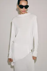 Jersey de punto liso blanco, con cuello redondo y mangas largas. Presenta un diseño de bajo asimétrico pronunciado.