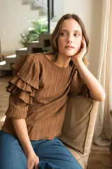 Blusa de tela texturizada color café, con cuello redondo y mangas al codo que presentan tres volados superpuestos y puños elásticos. Cuenta con cierre de botón en la parte posterior.