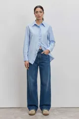 Camisa de algodón color celeste con cuello clásico y detalle de volados verticales a lo largo de la botonadura frontal. Presenta mangas largas con puños abotonados y un corte recto.