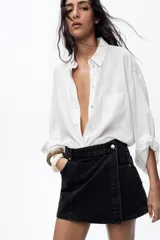 Falda pantalón de tiro alto confeccionada en denim negro. Presenta bolsillos delanteros y traseros de parche. El cierre frontal es cruzado con cremallera oculta y botón metálico.