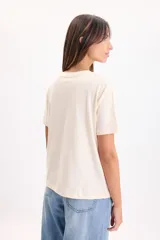 Remera manga corta de color crudo, con cuello redondo y estampa frontal ilustrada de Alicia en el País de las Maravillas rodeada de flores.
