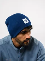 Gorro tipo beanie de tejido de punto acanalado en color verde oliva, con parche frontal rectangular blanco que presenta el logo de Umbro en rojo.