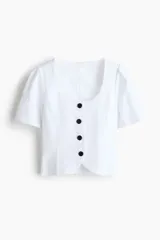 Blusa corta blanca con escote en U, mangas cortas abullonadas y cierre frontal con botones negros contrastantes.