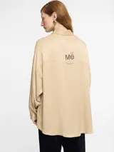 Camisa color beige de tejido satinado, corte oversize, manga larga, tapeta con botones invisibles y logo bordado en la espalda.