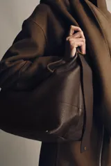 Bolso tipo saco de gran tamaño en cuero marrón oscuro, con textura granulada. Presenta un asa de hombro ancha y detalles metálicos dorados en la unión del asa. Incluye un bolsillo interior extraíble y cierre mediante botón imantado.