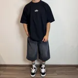 Remera negra de corte oversize con estampa rectangular en la espalda que representa una mano alcanzando un círculo sobre un fondo de líneas horizontales.