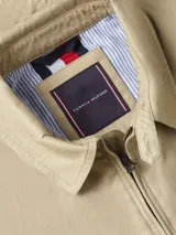 Campera tipo Harrington de algodón color beige claro, con cuello camisero y cierre frontal de cremallera. Presenta bolsillos laterales ribeteados y el logo de Tommy Hilfiger bordado en el pecho.
