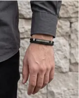 Pulsera doble de color negro, compuesta por una banda de cuerina trenzada y una placa metálica central con diseño de guarda griega grabada.