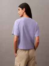 Remera lila de corte cropped y boxy, con cuello redondo y bolsillo en el pecho con escudo bordado en negro.