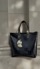 Bolso tipo tote bag negro con textura símil cuero de cocodrilo. Tiene doble asa corta, cierre superior y un dije colgante con forma de ala.