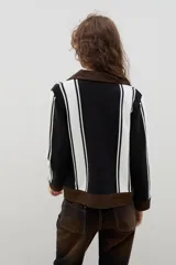 Campera de estilo casual confeccionada en una combinación de tejido de punto a rayas verticales en blanco y negro, con detalles en denim marrón en cuello, puños, ruedo y cierre frontal.