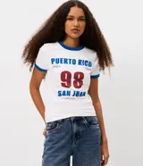 Remera de algodón con cuello redondo y mangas cortas, con vivos en color azul en cuello y mangas. Presenta una estampa frontal con la inscripción 'Puerto Rico 98 San Juan' en colores azul y bordó.