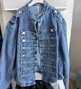 Chaqueta de denim estilo militar con cuello mao, cierre frontal con botones metálicos y detalles de trabillas horizontales. Presenta hombreras decorativas y diseño estructurado.