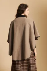 Poncho de tejido suave color beige con diseño envolvente y terminaciones con costuras decorativas en contraste color negro. Presenta un detalle de lazo frontal integrado.
