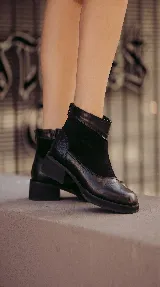 Bota corta de cuero negro con diseño de paneles que combina cuero liso y gamuza. Presenta puntera cuadrada, cierre trasero y suela con plataforma y taco bajo geométrico.