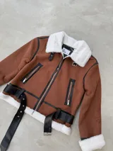 Campera estilo aviador en color camel, confeccionada en gamuza sintética con interior y cuello de piel sintética blanca. Presenta detalles en cuero sintético negro, cierre frontal, bolsillos laterales con cierre y cinturón ajustable en la cintura.