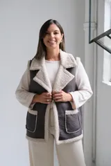 Chaleco de gamuza sintética en color gris oscuro con interior y bordes de corderito beige. Presenta solapas amplias, dos bolsillos frontales con ribetes de corderito y diseño sin cierre.