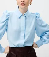 Camisa de crepé color blanco, con cuello inglés, manga larga abullonada con puños abotonados y cierre frontal oculto con botón diferenciado en el cuello.