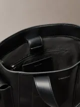 Cartera tote estructurada Calvin Klein de piel sintética lisa en color negro, con asas superiores de mano, compartimentos exteriores en los laterales y logo metálico grabado en la parte delantera.