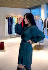 Vestido corto de satén color verde esmeralda, con mangas largas abullonadas, cuello redondo y diseño con drapeado lateral que genera un efecto asimétrico en el ruedo.
