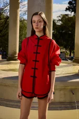 Camisa de algodón color rojo con ribetes negros, cuello mao y cierre frontal con nudos de estilo oriental, mangas abullonadas con puños ajustados.
