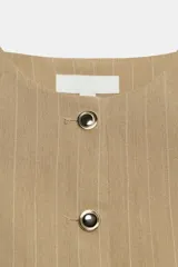 Chaleco corto de color beige con diseño de rayas sutiles, cuello redondo, sin mangas y cierre frontal con botones metálicos dorados. Presenta falsos bolsillos ribeteados en el delantero.
