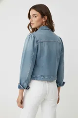 Chaqueta de jean negra con cuello camisero, cierre frontal con botones metálicos y mangas con sutiles frunces en los hombros. Presenta un corte entallado al cuerpo y bolsillos frontales con solapa.