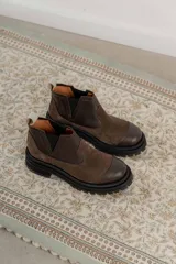Bota tipo chelsea de punta redonda, confeccionada en una combinación de cuero texturizado y gamuza en tonos marrones. Presenta elásticos laterales negros y una suela track robusta de goma negra.