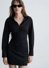 Vestido corto de estilo camisero, color negro con estampado de rayas verticales finas. Presenta cierre frontal con cremallera, detalle de fruncido central, cuello camisero y mangas largas con puños abotonados.