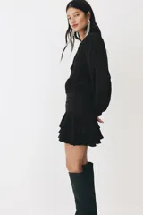 Vestido corto negro de mezcla de viscosa, con escote redondo, abertura frontal con tiras finas y mangas largas abullonadas con puños elásticos. Presenta sección entallada en la cadera con alforzas y falda corta con volados escalonados.