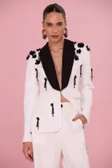 Blazer blanco de sastrería femenina con solapas negras de estilo esmoquin, bordados florales en relieve y detalles de pedrería en hombros y cuerpo.