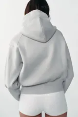 Chaqueta de punto con capucha, manga larga y cierre frontal con cremallera. Presenta un diseño de lazo decorativo en color amarillo en la zona del cuello.