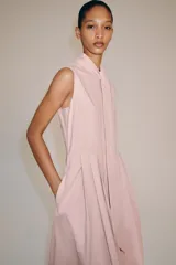 Vestido midi de Zara Woman Collection color rosado, confeccionado en hilatura de algodón. Presenta cuello subido con lazada, diseño sin mangas, bolsillos laterales ocultos y pliegues delanteros. Su falda tiene acabado en línea evasé y cuenta con cierre frontal mediante botones ocultos por solapa.